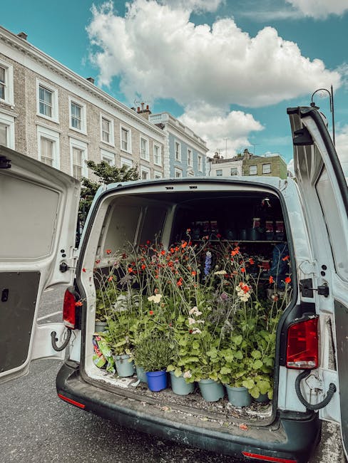 The open rear of a white delivery van filled with potted plants and flower stems, including red poppies and white blooms, arranged for flower delivery. The plants are in various blue and gray pots, wi