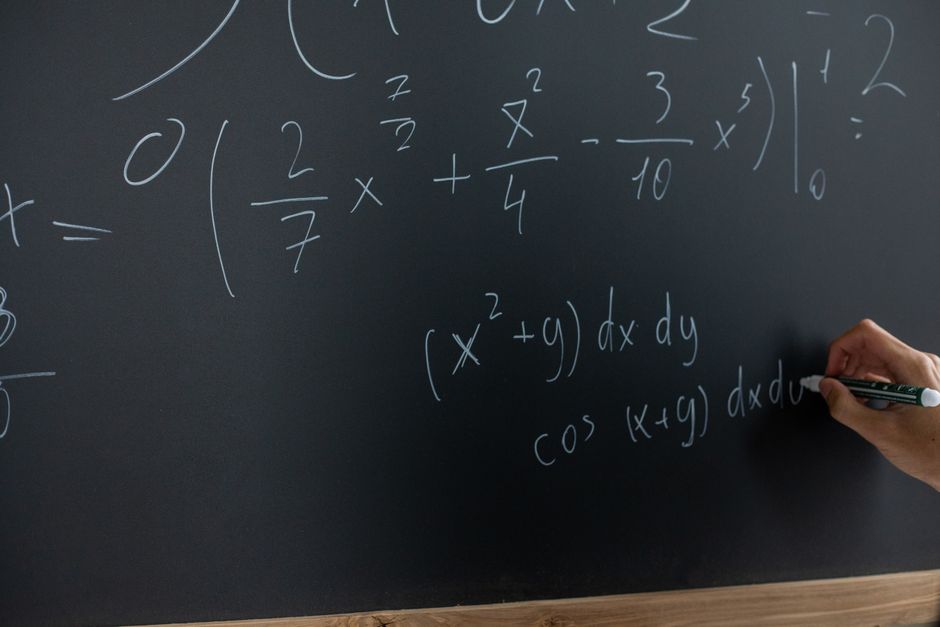 Photograph of a black chalkboard featuring handwritten mathematical equations and formulas in white chalk, with a person's hand in the lower right corner holding a black and white marker. The equation
