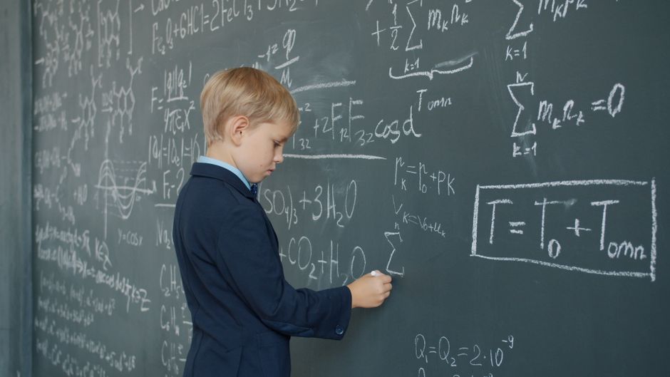 A young boy dressed in a navy blazer and light blue shirt is standing in front of a large chalkboard filled with complex mathematical equations and formulas written in white chalk. He is holding a pie