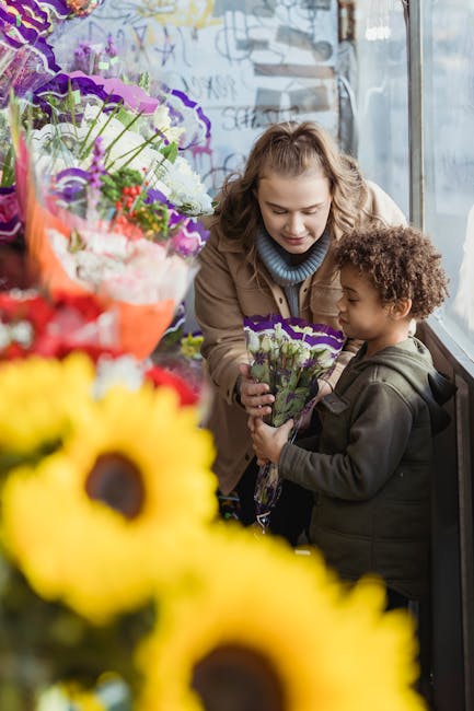 A young woman and a child stand inside a flower shop, closely examining a bouquet of purple and white flowers held by the woman. The arrangement features fresh, vibrant blossoms with delicate petals, 
