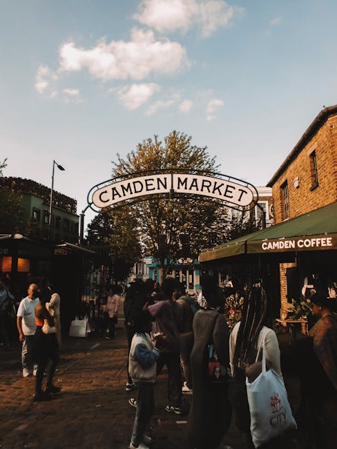 A vibrant bouquet of fresh flowers arranged with a mix of pink, purple, and white blooms, including roses and chrysanthemums, is held by a hand in front of Camden Market. The flowers are wrapped in cl